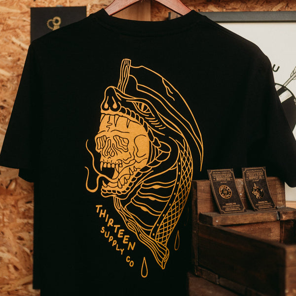 Black t-shirt with gold graphic design and 'Thirteen Supply Co' text on a wooden surface.